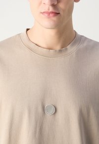 Beige puuvillane T-särk ümmarguse kaelusega ja halli värvi tekstuuriga ringikujuline logo padi rinna keskosas. Sujuv pind.