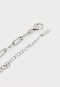 Chaîne en métal argenté avec des maillons allongés et un fermoir. Comprend une chaîne à maillons plus courte avec une étiquette rectangulaire gravée "LACOSTE."