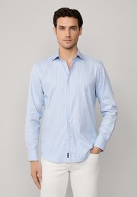 Camisa de manga larga a rayas en azul claro, hecha de algodón. Presenta un cuello abotonado y una pequeña etiqueta en el dobladillo. Se lleva con pantalones blancos.