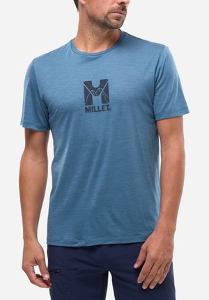 Homme portant un t-shirt à manches courtes bleu avec un logo "Millet" noir et un pantalon bleu marine, debout devant un fond clair uni.