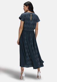 Lang donkergroene jurk met kapmouwen, voorzien van een patroon aan de achterkant, een tailleband en een sleutelgatdetail bij de halslijn. Gepaard met witte sandalen met bandjes.