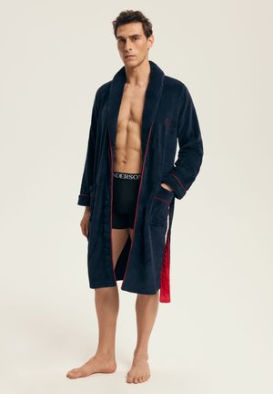 Mann, der einen marineblauen Bademantel mit roter Einfassung über schwarzen Boxershorts trägt, steht barfuß vor einem schlichten hellen Hintergrund.