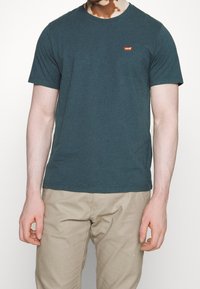 Mørk teal bomuld t-shirt med rund hals, korte ærmer og et lille rødt Levi's logo patch på brystet. Båret med lysebeige bukser.
