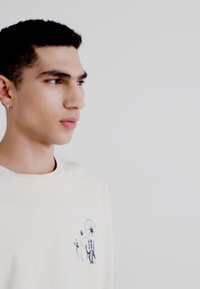 Tricou crem, cu mânecă lungă, cu un design grafic albastru care prezintă figuri abstracte și un emblema circulară în zona stânga a pieptului.