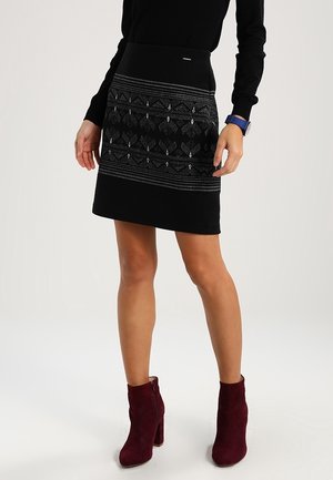 Schwarzer Rock mit einem strukturierten Taillenbund und geometrischen silbernen Mustern, kombiniert mit burgunderroten Wildleder-Stiefeletten mit Blockabsatz.