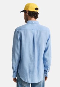 Camisa de botones azul claro con mangas largas, cuello y dobladillo redondeado. El material tiene un acabado texturizado. La parte posterior presenta un detalle de yugo único.