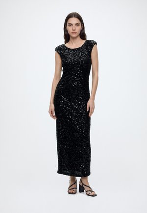Femme debout portant une robe longue noire sans manches à sequins et des sandales noires à talons blocs à brides sur un fond blanc uni.