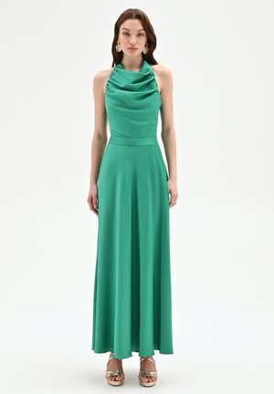 Donna che indossa un abito verde in raso senza maniche con scollo drappeggiato e sandali dorati con cinturini sottili, in piedi su sfondo bianco.