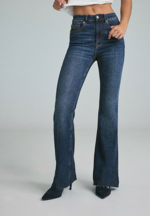 Personne portant un jean flare bleu foncé taille haute avec des ourlets effilochés et des escarpins noirs à bout pointu sur un fond gris clair.