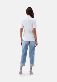 Weißes kurzärmliges Hemd, hellblaue, cropped Jeanshose und blaue High Heels. Das Hemd hat einen geraden Schnitt und aufgerollte Ärmel.