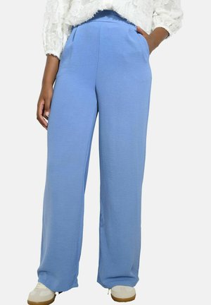 Persona che indossa pantaloni larghi azzurri chiari, una maglietta bianca con texture e sneakers bianche, con una mano in tasca.
