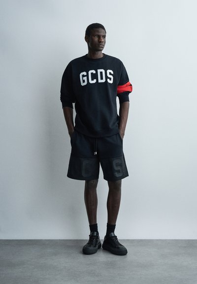 Άνδρας στέκεται με τα χέρια στις τσέπες φορώντας μαύρο φούτερ GCDS με κόκκινο περιβραχιόνιο, ταιριαστά μαύρα σορτς, κάλτσες και αθλητικά παπούτσια μπροστά σε απλή λευκή επιφάνεια.