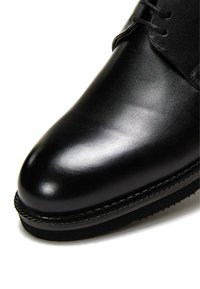 Zapato de cuero negro con acabado brillante, puntera redondeada y cierre de cordones. Suela de goma con detalle de costura a lo largo del borde.