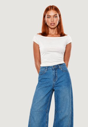 Donna con capelli rossi che indossa un top bianco scollato sulle spalle e jeans blu a gamba larga a vita alta, in piedi con le mani dietro la schiena.