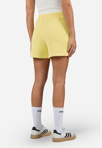 Pantaloni corti gialli a coste con una vita elastica, di lunghezza fino a metà coscia e con una superficie liscia. Indossati con calzini bianchi e sneakers.
