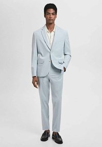 Jasnoniebieski garnitur o dopasowanym kroju, z marynarką zapinaną na dwa guziki, wyłogami i prostymi spodniami. Noszony z kremową koszulą i czarnymi butami.