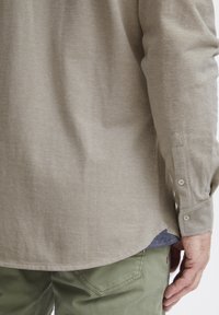 Chemise longue à manches longues de couleur brun clair avec des poignets à boutons. L'ourlet a une forme courbée. En dessous, une chemise bleue est visible au niveau du col.
