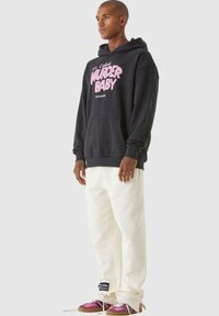 Felpa nera con testo rosa che recita "Si chiama OMICIDIO PICCOLA" e tasca frontale. Pantaloni bianchi, sneaker rosa e marroni con dettagli neri.