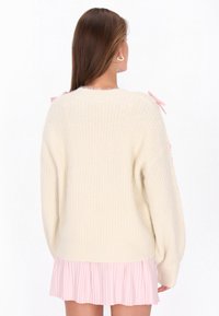 Cremfarbener Strickpullover mit übergroßen Ärmeln, der an den Schultern mit rosa Schleifen verziert ist, sowie einem rosa plissierten Rock am Saum. Weiche Textur.