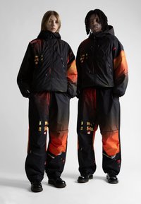 Melnas ārējās apģērba detaļas ar oranžām gradienta akcentiem un abstraktiem rakstiem, ar rāvējslēdzējiem un kabatām, ko valkā divi atēli, stāvot plecu pie pleca.