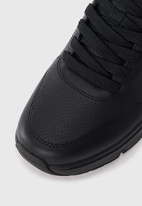Chaussure de sport noire avec une tige en cuir texturé, bout rond et lacets plats noirs. Des détails perforés ajoutent de la respirabilité au design.