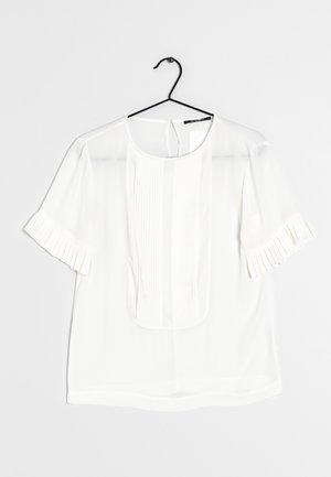 Biała bluzka z krótkim rękawem, z plisowanym detalem na przednim panelu i mankietach rękawów, zawieszona na czarnym wieszaku na białym tle.