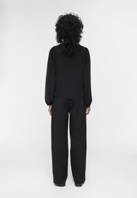 Blouse noire à manches bouffantes, associée à un pantalon noir ample. Le tissu semble lisse et l'ensemble présente un design monochrome.
