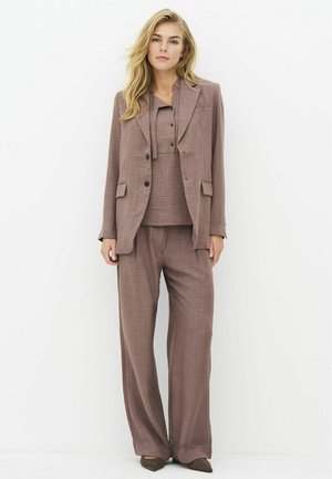 Woman standing wearing a loose brown suit with wide-leg pants, a matching buttoned shirt, and a blazer against a plain white background.