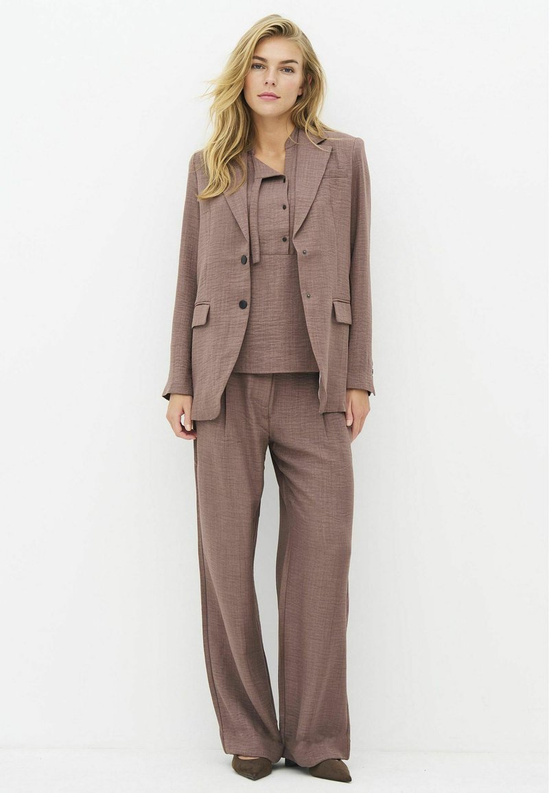Femme debout portant un costume marron ample avec un pantalon à jambes larges, une chemise boutonnée assortie et un blazer sur un fond blanc uni.