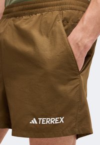Shorts marron avec ceinture élastique et poche latérale, une main dans la poche et logo blanc "TERREX" près de l'ourlet.