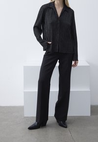 Černá kovová košile s dlouhými rukávy a límcem, spárovaná s černými kalhotami s vysokým pasem a černými botami na podpatku s špičatou špičkou.