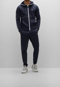 Mörkblå hoodie med dragkedja framtill, justerbara snoddar och sidostripes; tillsammans med matchande joggers och ljusa sneakers.