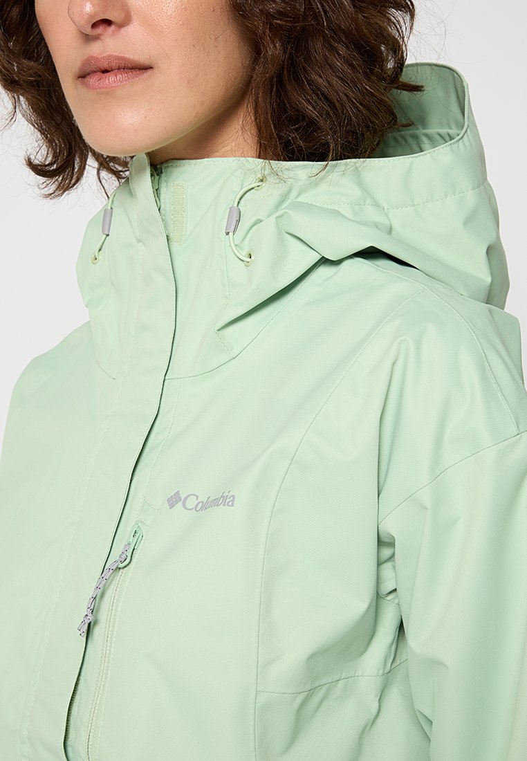Veste Columbia vert clair avec zip sur le devant, capuche réglable et tissu lisse résistant à l'eau, avec un logo discret sur la poitrine.