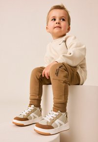 Sweatshirt crème, joggers marron avec logo sur le côté, et baskets grises avec accents marron. Matériaux lisses, coupe décontractée et design superposé.