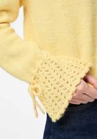 Żółty dziergany sweter z teksturowanym koronkowym rękawem o falistym brzegu i wiązanej dekoracji. Miękka, lekka tkanina odpowiednia do nakładania warstw.