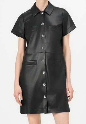 Krótka czarna skórzana sukienka z przodu na srebrne guziki, z krótkimi rękawami, kołnierzykiem i czterema klapowymi kieszeniami, noszona przez stojącą osobę.