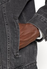 Grå denimjacka med synlig sömnad, en ficka och en texturerad yta. En hand är placerad i fickan och visar hudton.