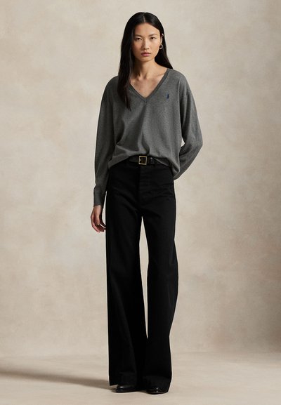 Moteris stovi, dėvi laisvą pilką V formos megztinį įkištą į juodas, plačiomis kelnėmis su aukštu liemeniu, su juodu diržu ir juodais batais.