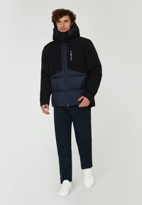 Svart och marinblå puffjacka med dragkedja, har en huva och två fickor med dragkedja, bärs med marinblå byxor och vita sneakers.