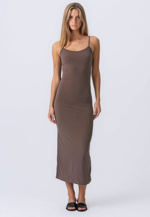 Egyenes hajú nő, barna, testhez simuló, ujjatlan maxi ruhában és fekete papucsban, világos, egyszerű háttér előtt áll.