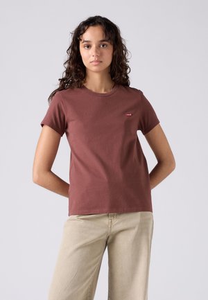 Fiatal nő göndör hajjal áll, kezeit a háta mögött tartva, barna rövid ujjú pólót és világos bézs nadrágot visel.