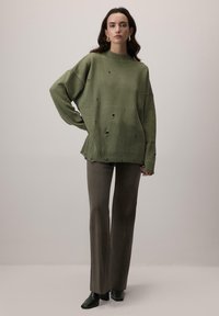 Przesadnie duży zielony sweter z wysokim kołnierzem i postrzępionymi dziurami, w zestawieniu z dopasowanymi ciemnymi spodniami i czarnymi, spiczastymi botkami.