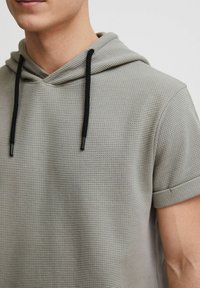 Sudadera de manga corta en gris, confeccionada en una tela texturizada con una sensación ligera. Presenta una capucha con cordón y detalles en negro, así como mangas enrolladas.