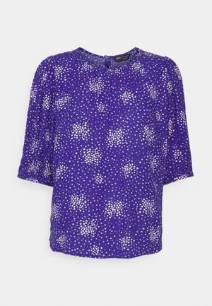 Fioletowa bluzka z krótkimi puffowanymi rękawami, z wzorem białych kropek i marszczonym dekoltem. Wykonana z lekkiego materiału.
