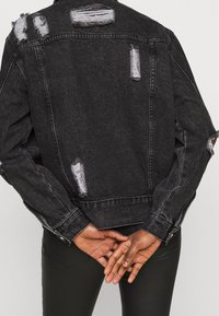 Osoba v černé džínové bundě s desgátem a roztrhanými detaily, s rukama spojenýma za zády, v kombinaci s černými kalhotami.