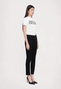 Λευκή μπλούζα με τύπωμα "Versace Jeans Couture" σε μαύρο χρώμα, συνδυασμένη με εφαρμοστά μαύρα τζιν και μυτερές μαύρες γόβες.