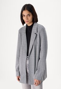 Grey patterned blazer with a relaxed fit, notched lapels, and long sleeves. Worn over a black top, paired with light-colored jeans.
