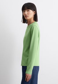 Lange mouwen groene katoenen t-shirt met een ronde halslijn. Model staat zijwaarts, waardoor een eenvoudig en slim ontwerp zichtbaar is. Donkerdenim jeans zichtbaar.