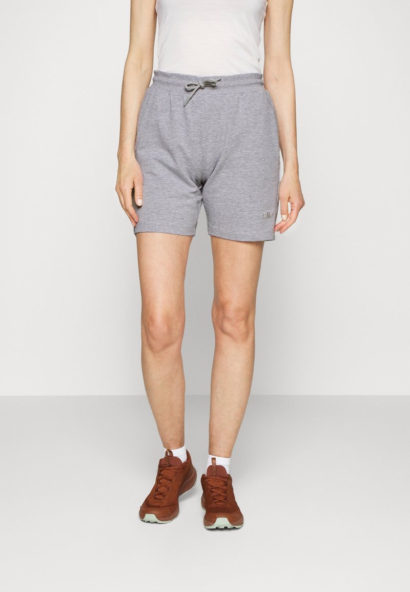 Femme portant un short gris à cordon, des chaussettes blanches, des chaussures de sport marron et un haut blanc sans manches, debout devant un fond uni.