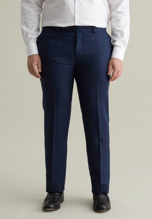 Pantalones de vestir navy hechos de un tejido de textura suave. Corte ajustado con pliegues en el frente, bolsillos laterales y cierre de botón. Llevados con zapatos negros.